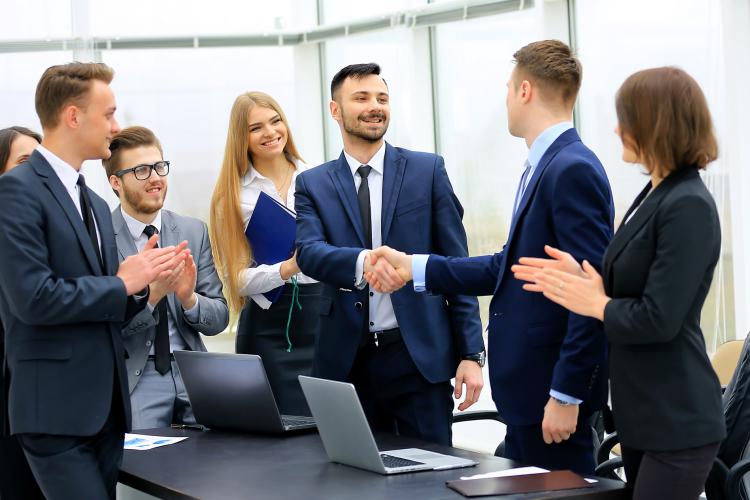 Group of confident business people in formal wear sitting at the table together and smiling while two men handshaking